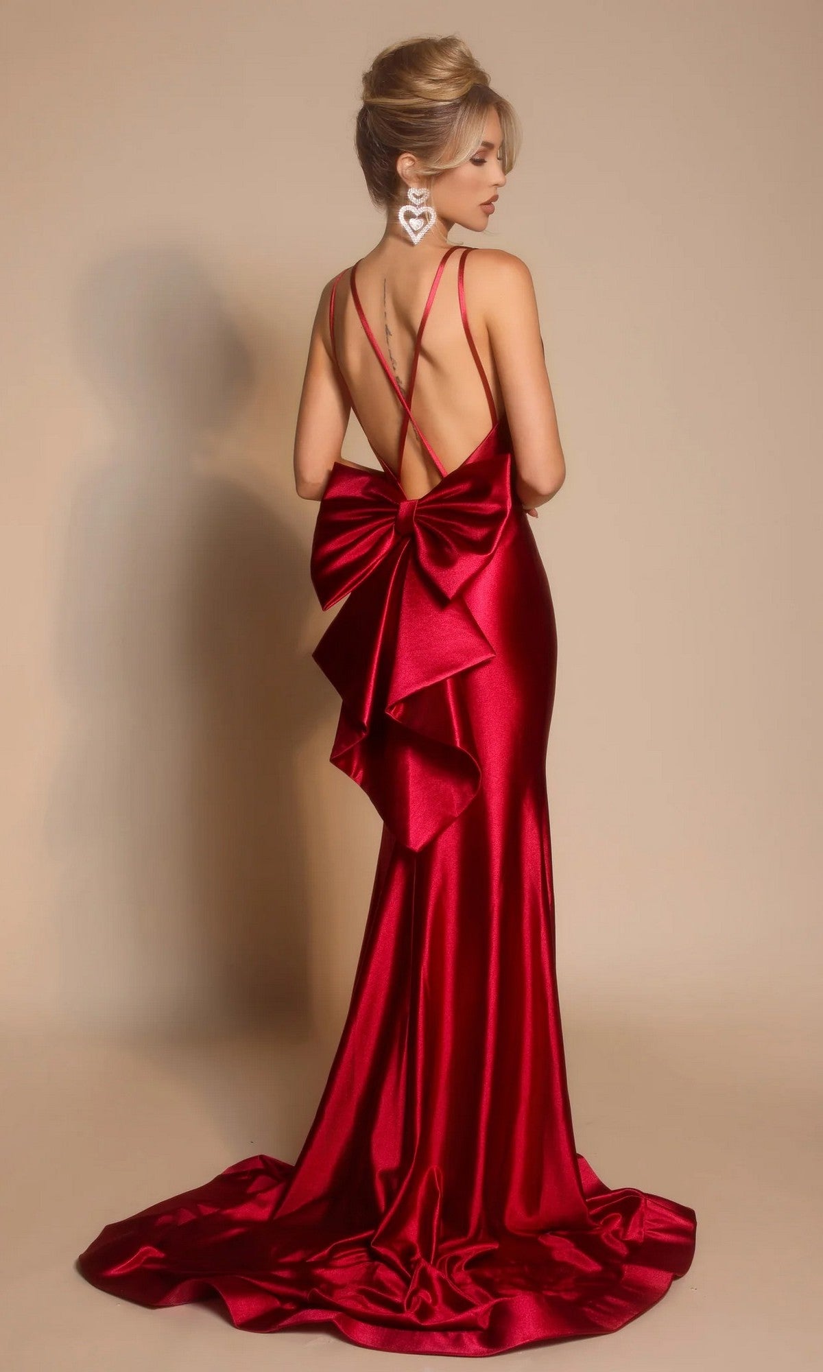A woman models the Portia and Scarlett XO095 long formal dress in burgundy, featuring a statement bow at the back, spaghetti straps, and a low open back, posed against a neutral background.